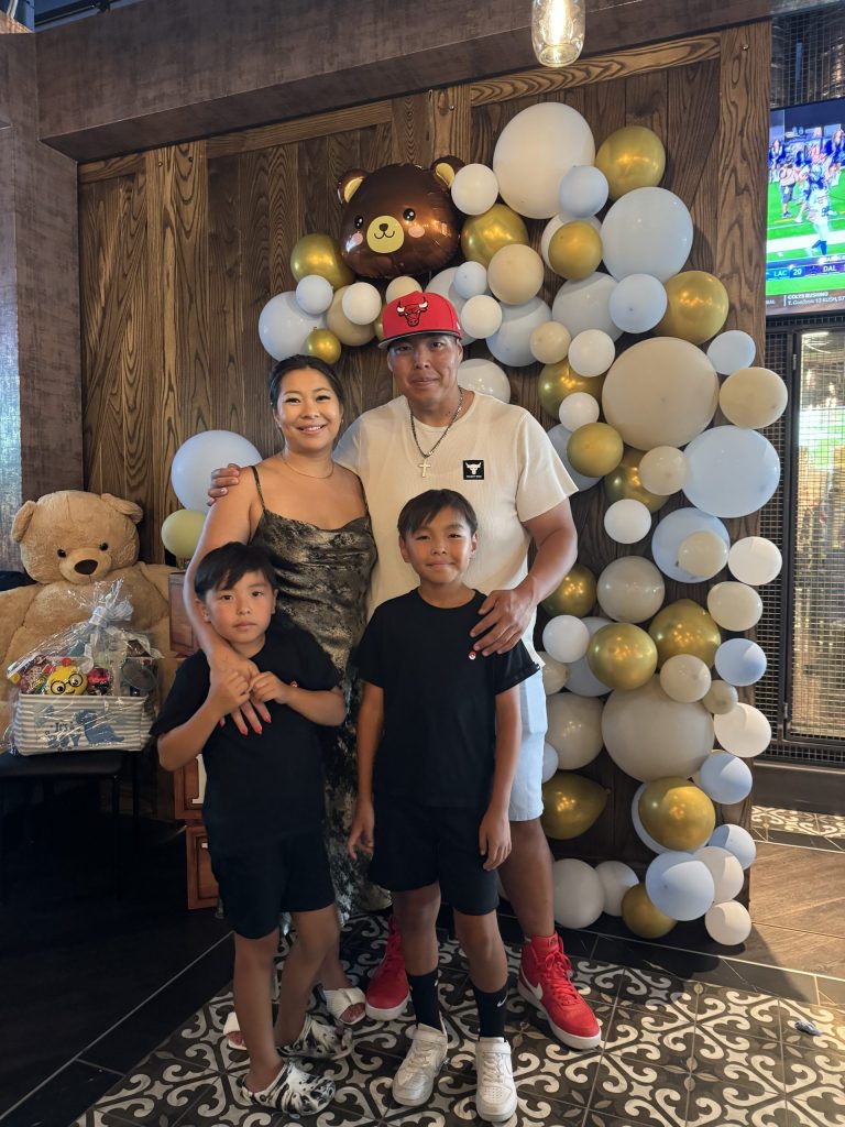 Bayar, Nikki, and their two children pose in front of a balloon arch at a celebration.